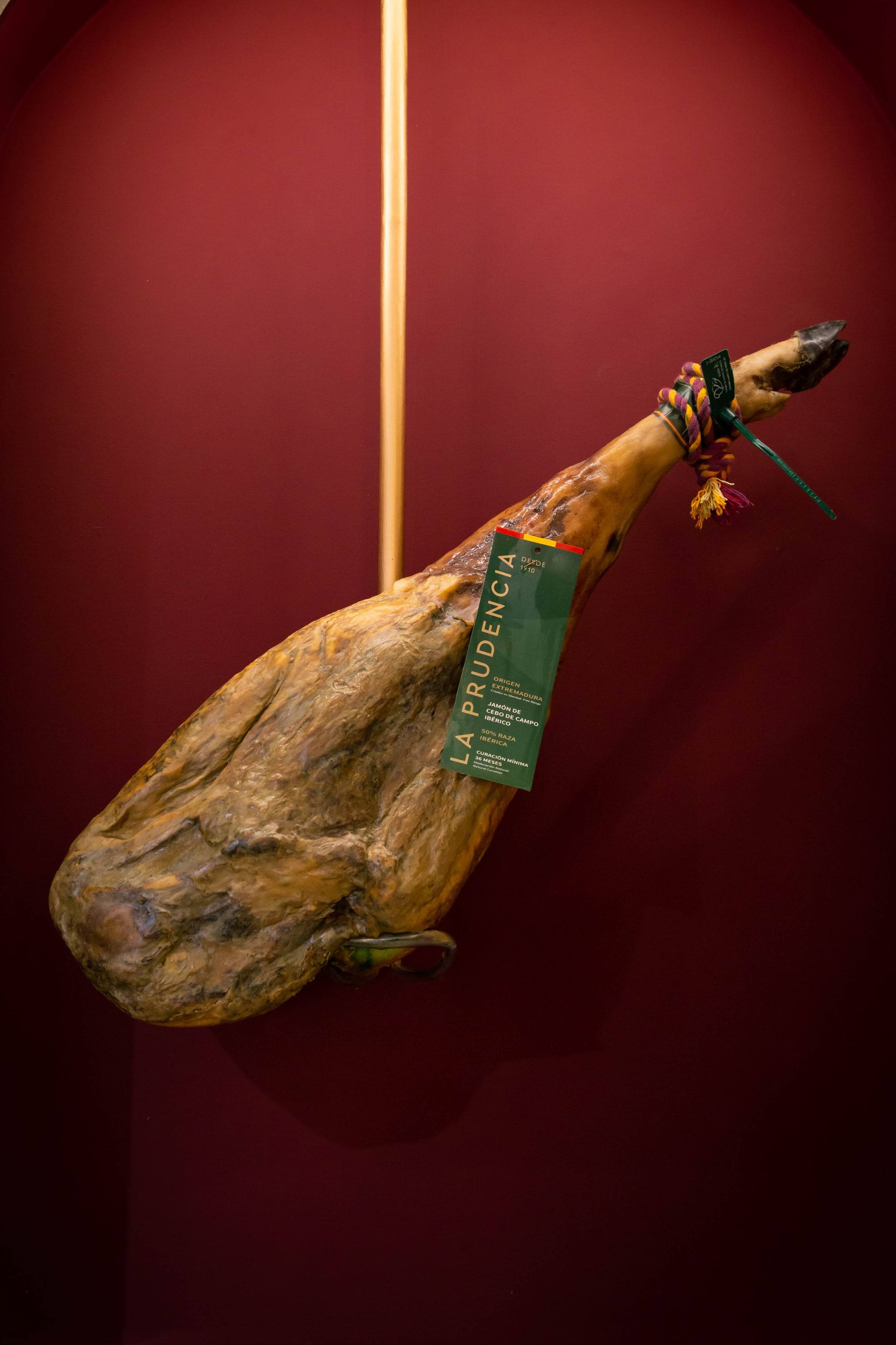 Jamón ibérico colgado con la etiqueta de La Prudencia sobre una pared granate.