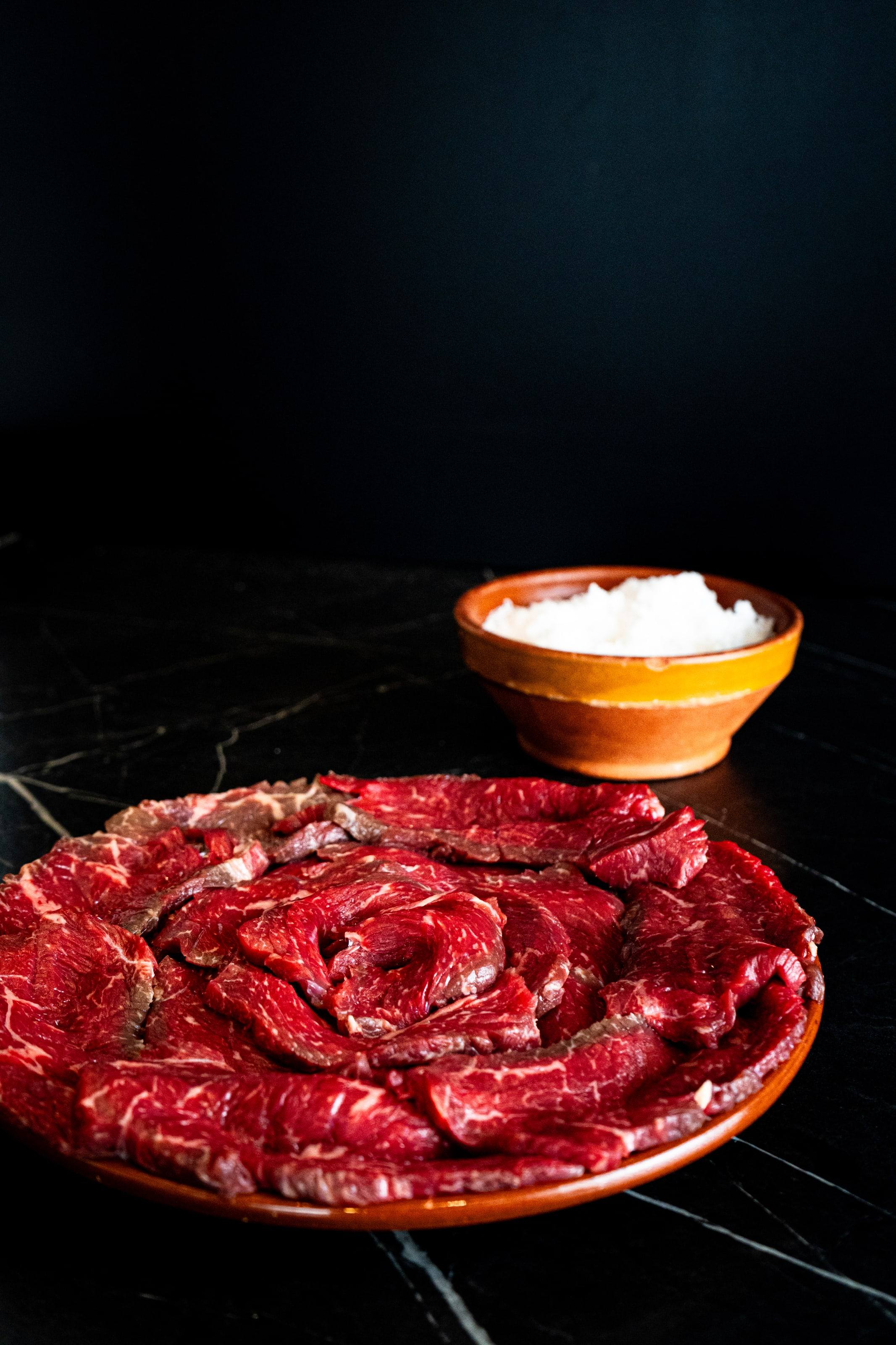 Plato de cecina en lonchas finas con un cuenco de sal sobre una mesa oscura.