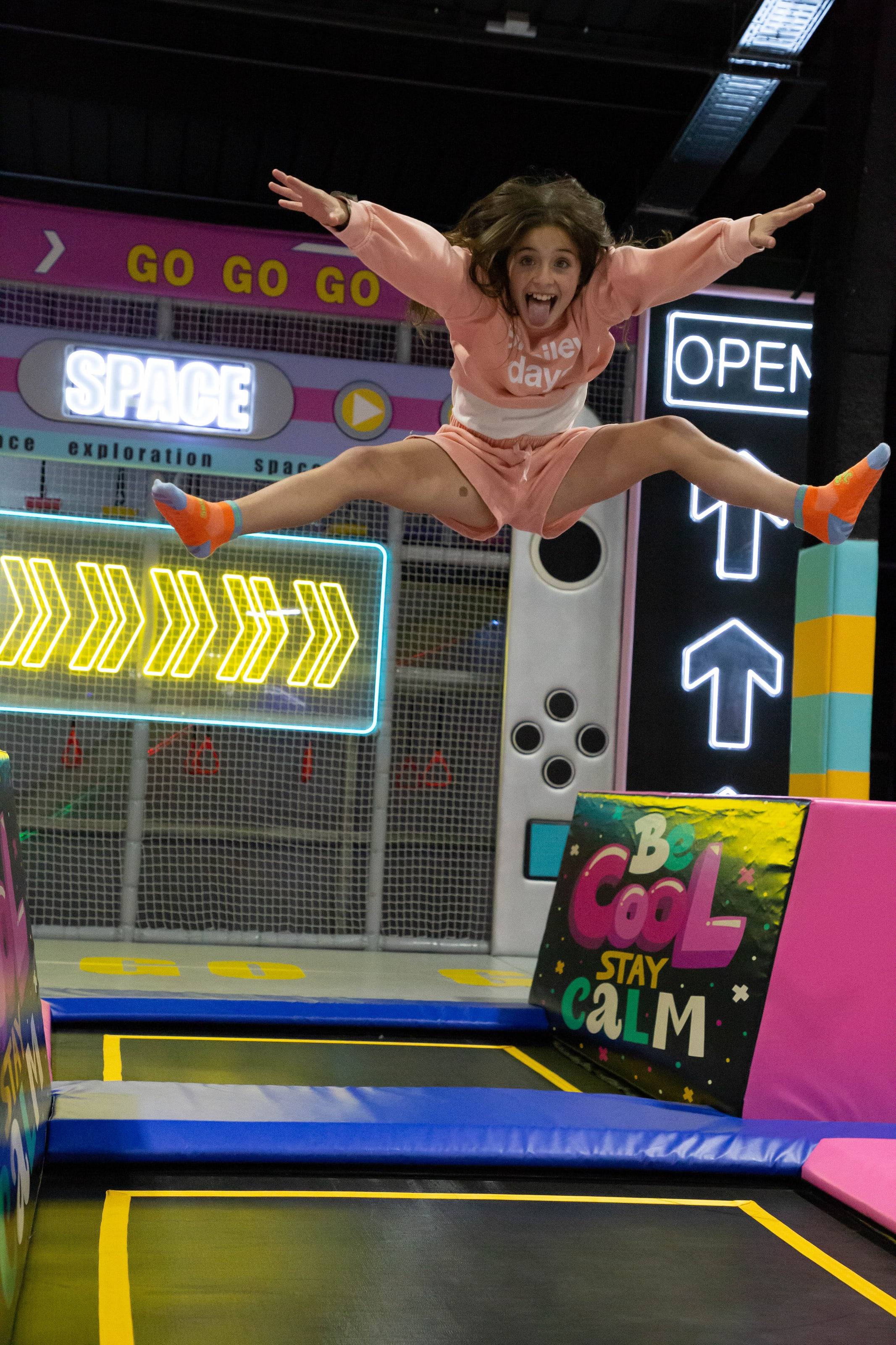 Niña saltando con las piernas abiertas en un parque de trampolines con luces de neón.